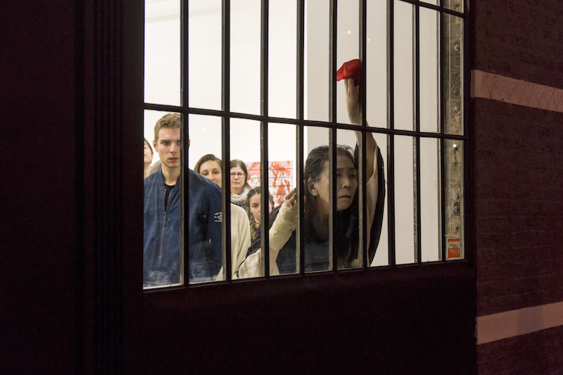 Eiko peers out into the street from behind a barred window. She looks defeated as audience members watch her in the background